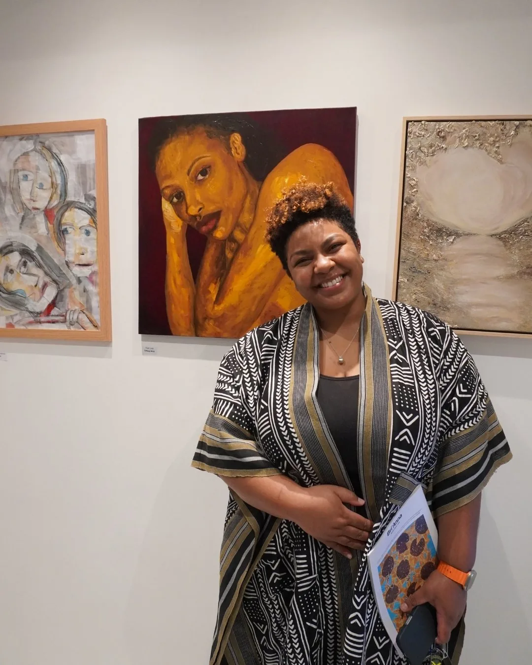 A smiling gallery visitor holding a magazine beside exhibited paintings.